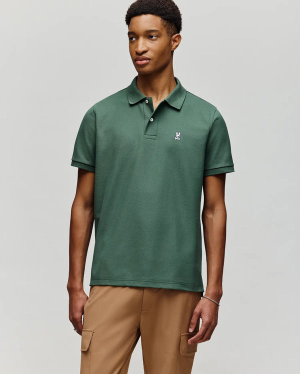 A young man wearing a Psycho Bunny MENS CLASSIC PIQUE POLO SHIRT - B6K001F200 in dark green with an embroidered Bunny logo and light brown cargo pants stands against a plain light gray background.
