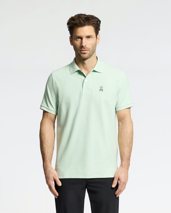 A man wears a Psycho Bunny Men's Classic Pique Polo (B6K001F200) in light green with an embroidered bunny logo on the chest, paired with black pants. He has short, styled hair and a neutral expression against a plain, light-colored background.