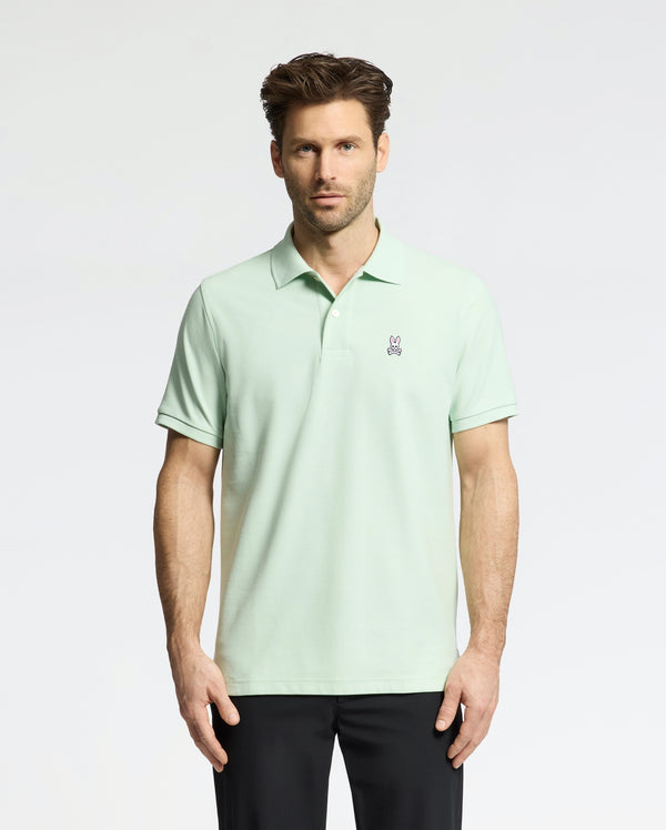 A man wears a Psycho Bunny Men's Classic Pique Polo (B6K001F200) in light green with an embroidered bunny logo on the chest, paired with black pants. He has short, styled hair and a neutral expression against a plain, light-colored background.