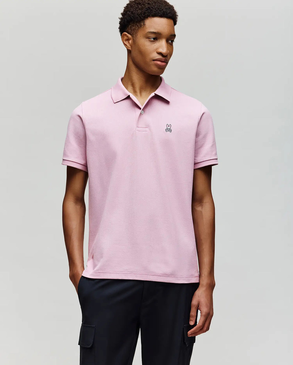 A young man stands against a plain background wearing Psycho Bunny’s MENS CLASSIC PIQUE POLO SHIRT - B6K001F200 in light pink, with an embroidered Bunny logo on the chest, paired with dark cargo pants. He looks to the side with one hand in his pocket.