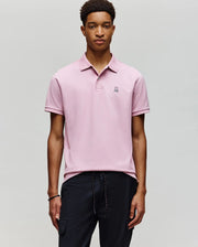 A young man wears a Psycho Bunny MENS CLASSIC PIQUE POLO SHIRT - B6K001F200 in light pink with an embroidered Bunny logo on the chest, paired with black drawstring pants, and stands against a plain light gray background.