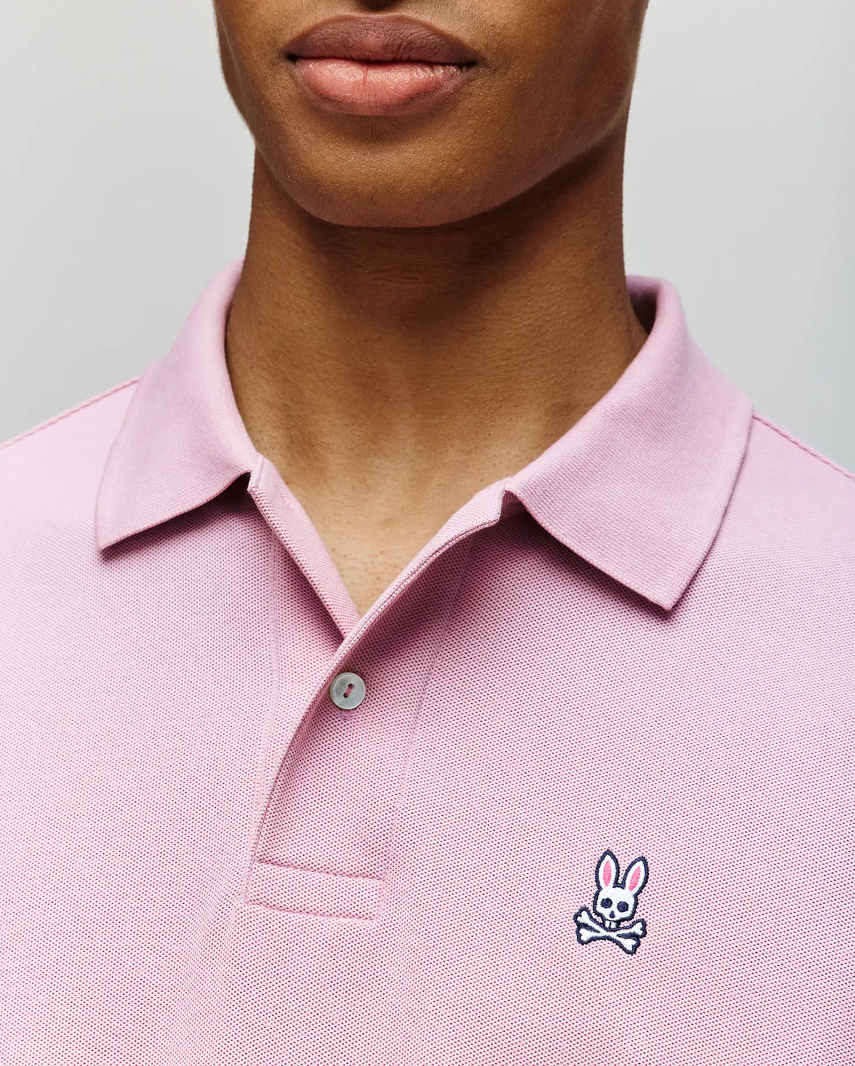 A person wears the Psycho Bunny MENS CLASSIC PIQUE POLO SHIRT (B6K001F200) in light pink, featuring a collared design, buttons, and the signature embroidered Bunny with crossbones logo on the chest. Only the lower face and upper torso are visible.