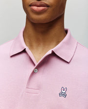 A person wears the Psycho Bunny MENS CLASSIC PIQUE POLO SHIRT (B6K001F200) in light pink, featuring a collared design, buttons, and the signature embroidered Bunny with crossbones logo on the chest. Only the lower face and upper torso are visible.