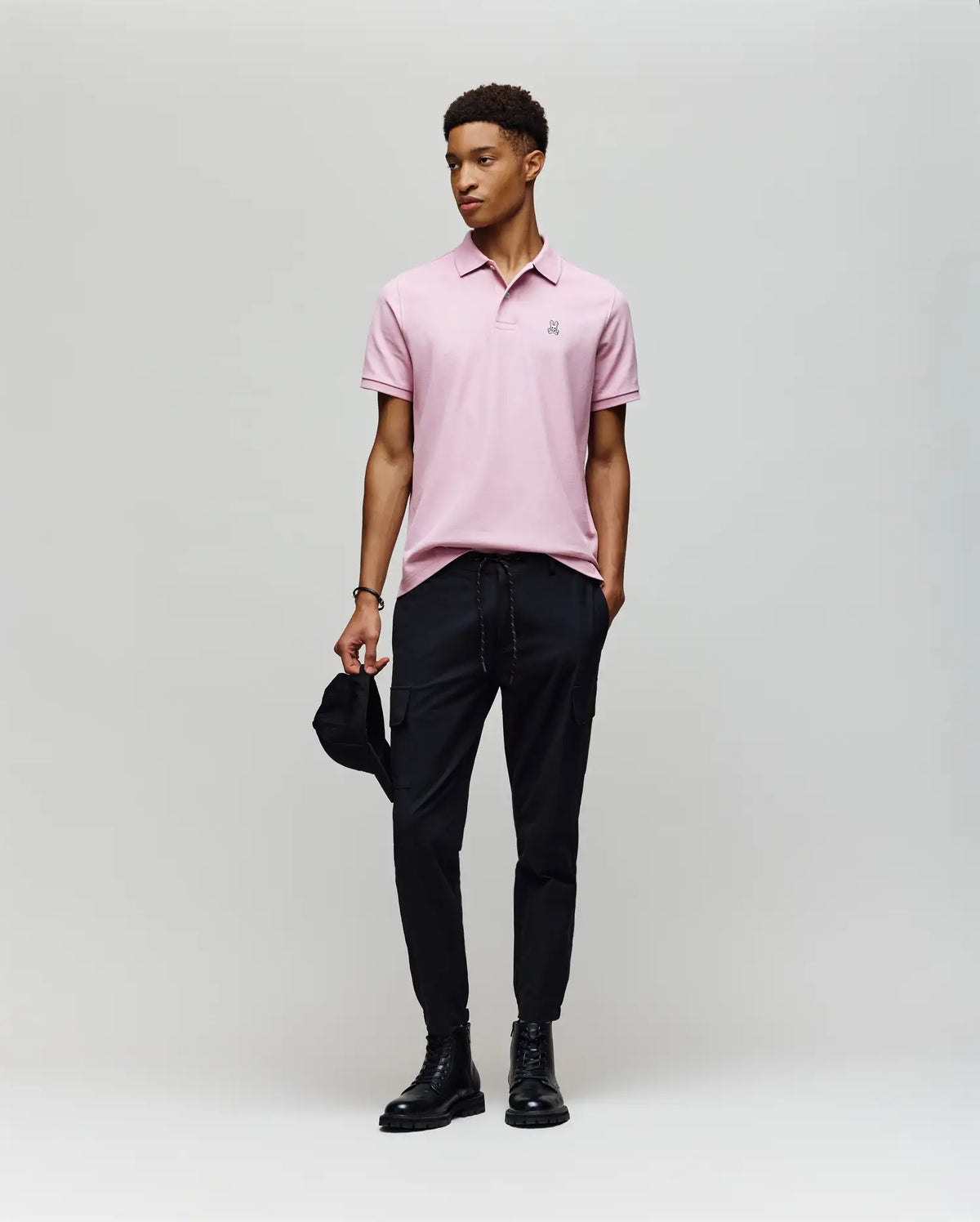 A man stands against a plain background wearing a Psycho Bunny MENS CLASSIC PIQUE POLO SHIRT (B6K001F200) in light pink with an embroidered Bunny logo, paired with black pants and boots. He holds a black cap and looks off to the side, appearing relaxed.