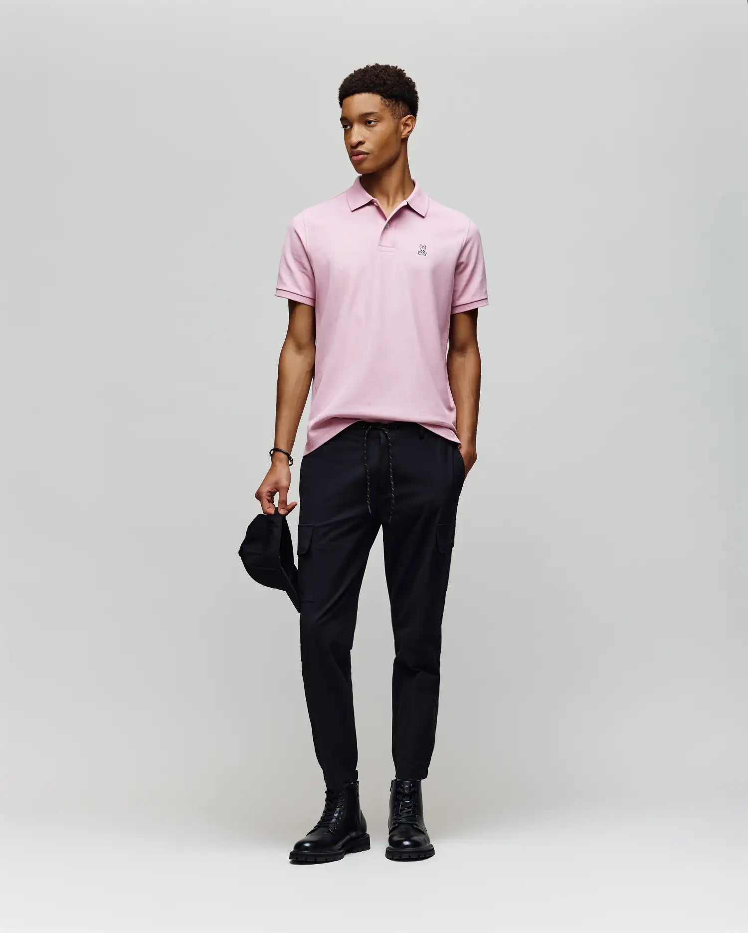 A man stands against a plain background wearing a Psycho Bunny MENS CLASSIC PIQUE POLO SHIRT (B6K001F200) in light pink with an embroidered Bunny logo, paired with black pants and boots. He holds a black cap and looks off to the side, appearing relaxed.