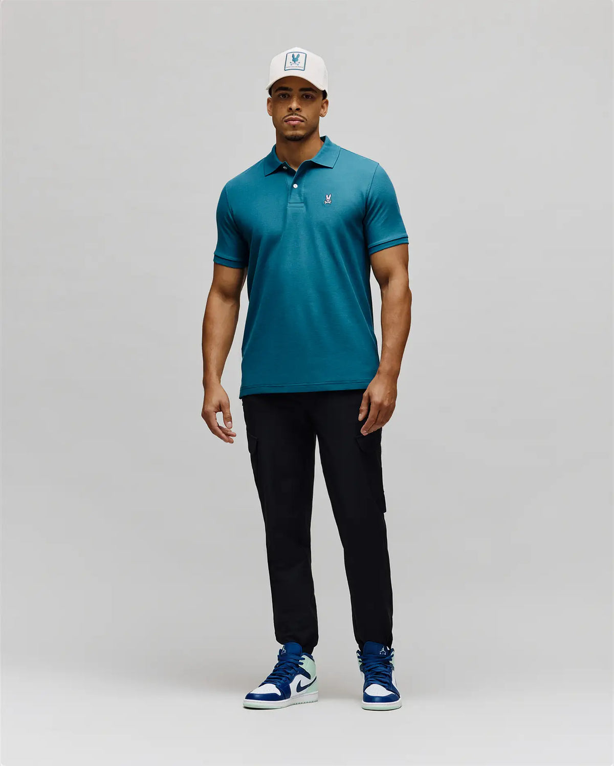 Mens Classic Pique Polo in teal, classic fit, with a white embroidered Psycho Bunny skull logo on the left chest. Paired with black cargo pants, a white logo cap, and blue, white, mint sneakers.