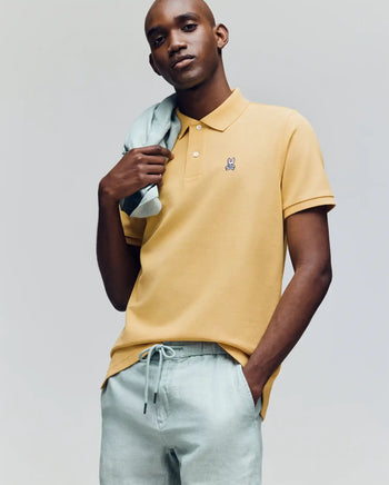 A man wears a Psycho Bunny MENS CLASSIC PIQUE POLO - B6K001F200 in yellow and light green drawstring pants, posing with a light green sweater over his shoulder against a plain background.