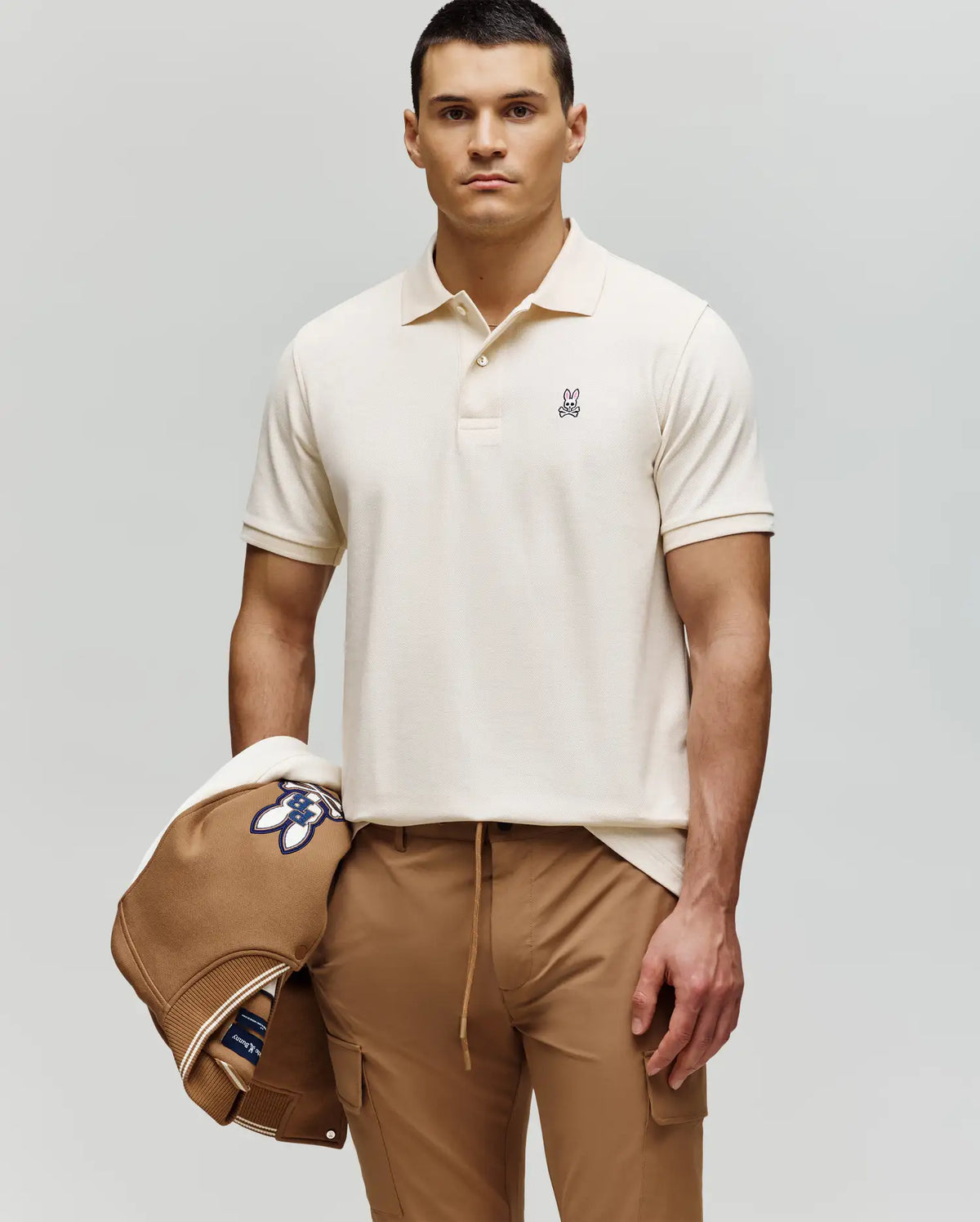 A man with short dark hair and a neutral expression wears the Psycho Bunny MENS CLASSIC PIQUE POLO SHIRT - B6K001F200 in cream and tan pants, holding a tan jacket with a blue and white patch against a plain light gray background.