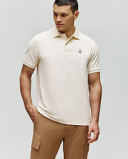 A man wears the Psycho Bunny MENS CLASSIC PIQUE POLO SHIRT (B6K001F200), featuring an embroidered Bunny logo, with tan cargo pants. He stands against a plain light background, looking down and slightly to the side.