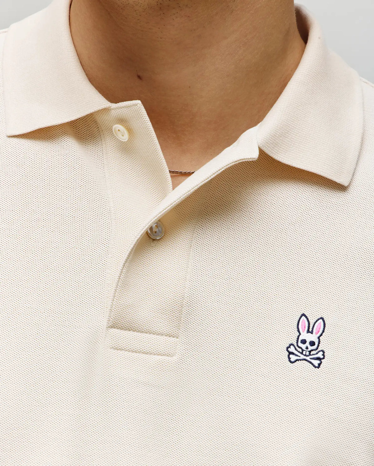 Close-up of a person wearing the Psycho Bunny MENS CLASSIC PIQUE POLO SHIRT - B6K001F200 in cream, with two buttons undone and a small embroidered Bunny logo with pink ears and crossbones on the left chest.
