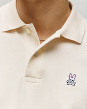 Close-up of a person wearing the Psycho Bunny MENS CLASSIC PIQUE POLO SHIRT - B6K001F200 in cream, with two buttons undone and a small embroidered Bunny logo with pink ears and crossbones on the left chest.
