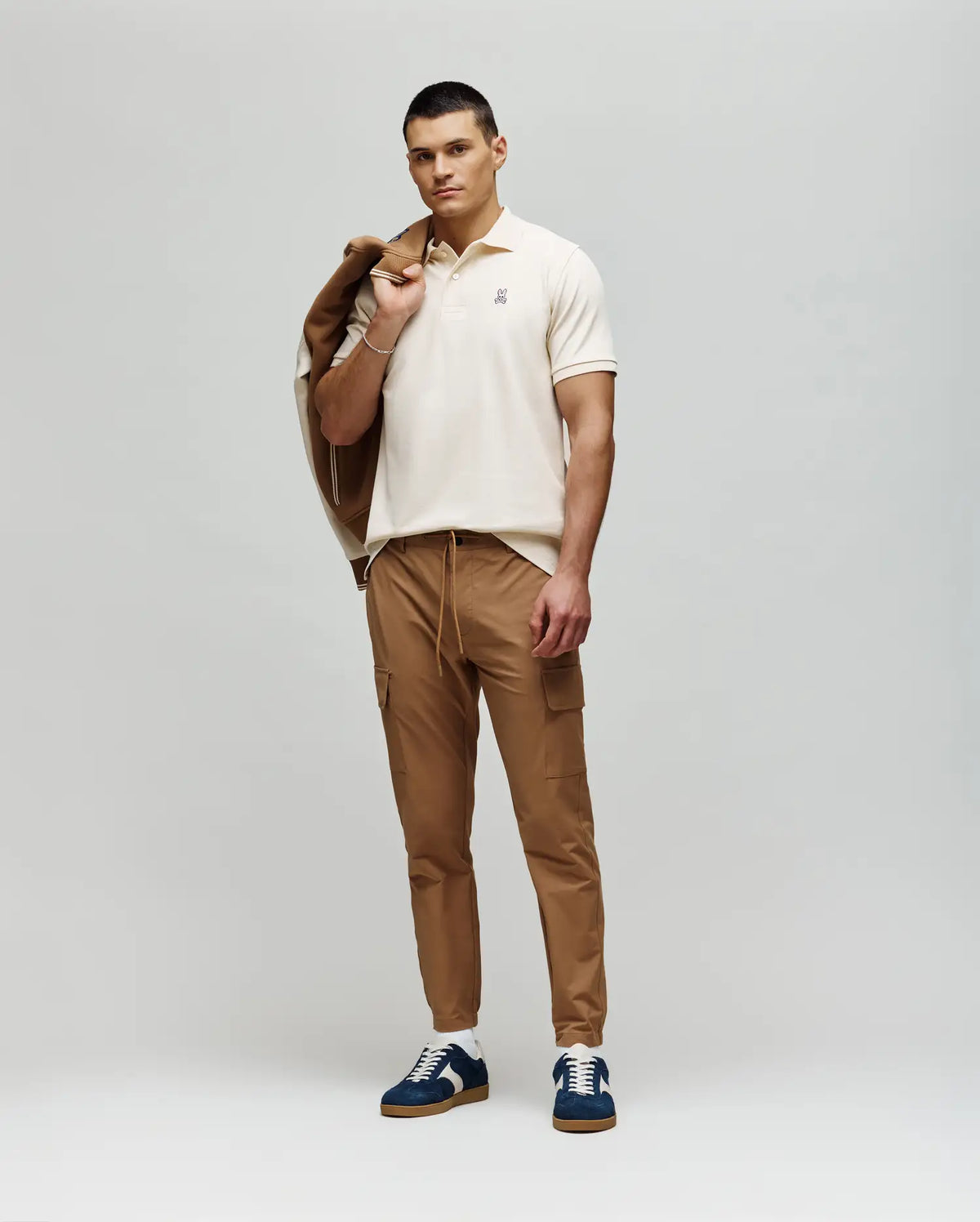 A man stands against a light background in a beige Psycho Bunny MENS CLASSIC PIQUE POLO SHIRT (B6K001F200), tan cargo pants, blue and white sneakers, holding a tan jacket over his shoulder.