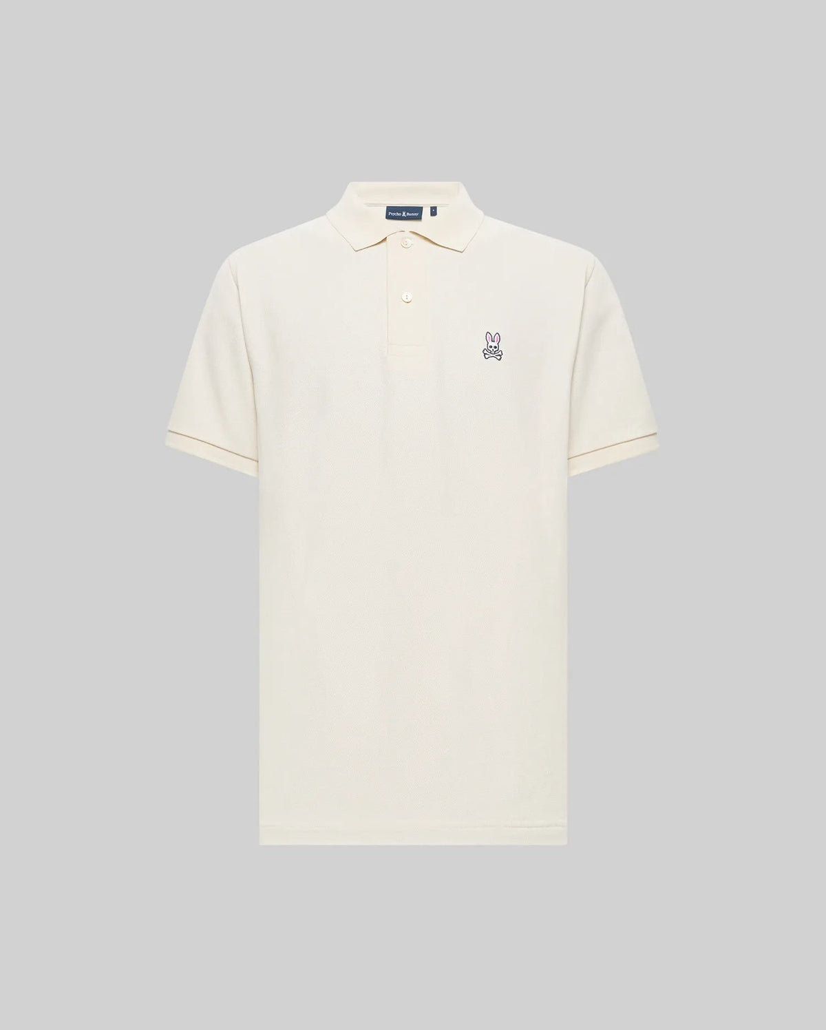 The Psycho Bunny MENS CLASSIC PIQUE POLO SHIRT (B6K001F200) is a cream-colored Pima cotton polo with a collar, button placket, and embroidered Bunny logo on the left chest, shown against a plain gray background.