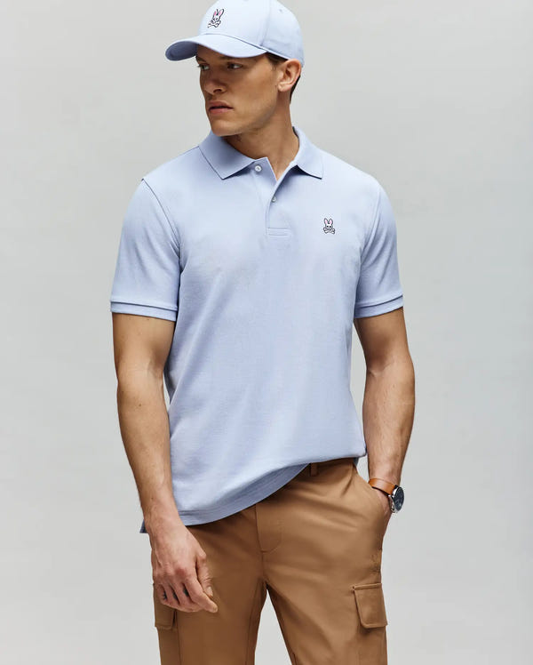A man stands against a plain background wearing a Psycho Bunny MENS CLASSIC PIQUE POLO SHIRT - B6K001F200 in light blue, an embroidered Bunny logo, a matching light blue baseball cap, tan cargo pants, and a watch.