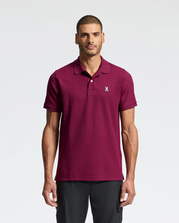 A man wearing a Psycho Bunny MENS CLASSIC PIQUE POLO SHIRT - B6K001Z1PC in burgundy stands against a plain light background. He has short hair and a neutral expression, with his hands relaxed at his sides, highlighting the soft elegance of Pima cotton and the embroidered Bunny logo.