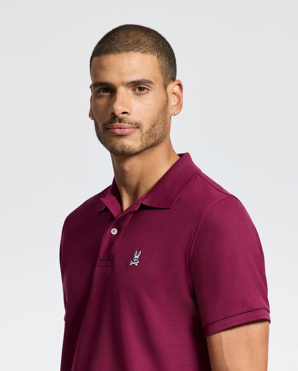 A man with short hair is wearing a burgundy MENS CLASSIC PIQUE POLO SHIRT - B6K001Z1PC from Psycho Bunny, made from soft Pima cotton and showcasing a small embroidered Bunny logo. The background is light grey.