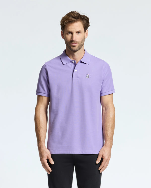 A man dressed in a Psycho Bunny light purple men's Classic pique polo (B6K001Z1PC), made from Pima cotton and adorned with a small embroidered Bunny logo on the chest, stands against a plain white background. He has short hair and pairs the polo with black pants.