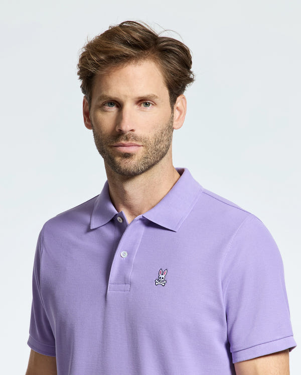 A man with light brown hair and a beard wears the Psycho Bunny MENS CLASSIC PIQUE POLO in lavender, showcasing an embroidered Bunny logo on the chest. The Pima cotton fabric adds to its comfort while he looks forward with a neutral expression against a plain background.