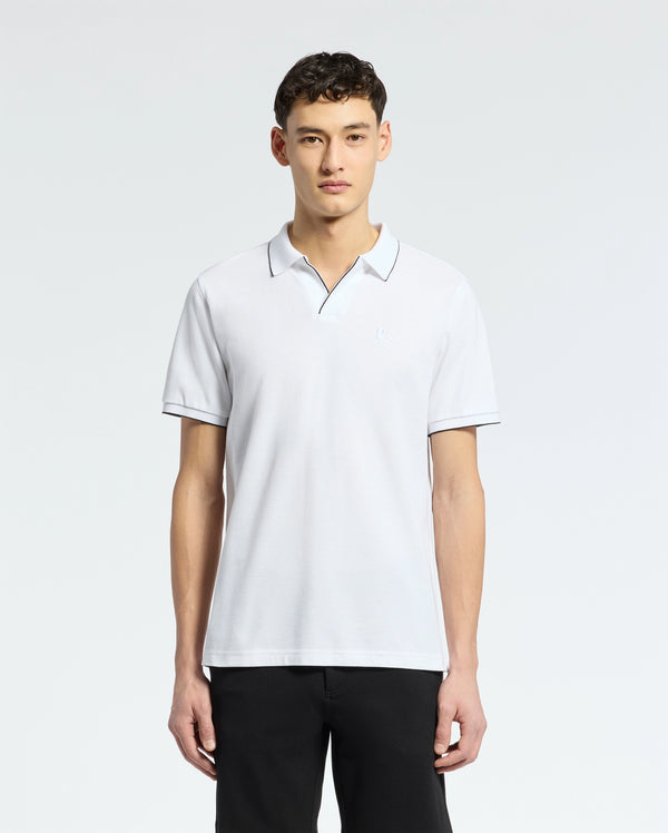 Wearing a Psycho Bunny Mens East Hills Johnny Collar Polo made of Pima cotton, the person stands against a white background. The white shirt features dark piping on the collar and a subtle chest logo. Their short, dark hair complements black pants.