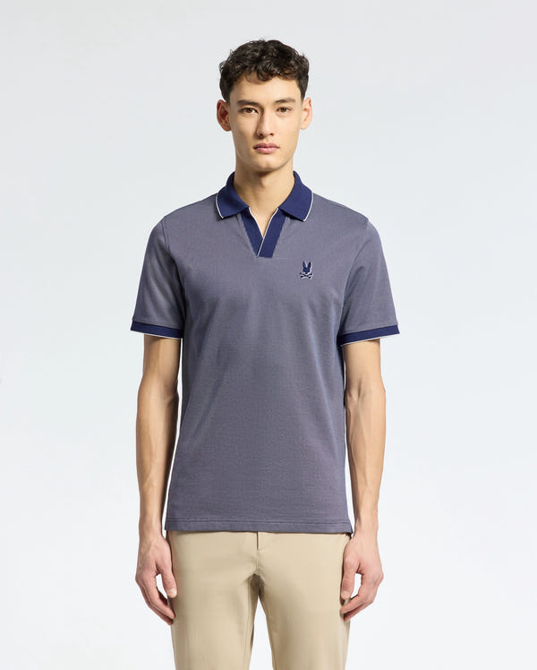A young man with short dark hair and a neutral expression wears Psycho Bunny's Mackay Jacquard Johnny Collar Polo (B6K408F200), a short-sleeved, blue Pima cotton shirt with a small chest logo, against a plain background and paired with beige pants.