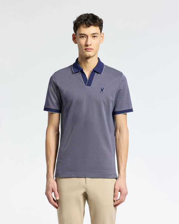 A young man with short dark hair and a neutral expression wears Psycho Bunny's Mackay Jacquard Johnny Collar Polo (B6K408F200), a short-sleeved, blue Pima cotton shirt with a small chest logo, against a plain background and paired with beige pants.