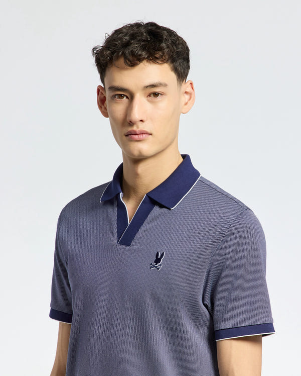 A person with short, dark hair wears a Psycho Bunny Men's Mackay Jacquard Johnny Collar Polo (B6K408F200) in blue, crafted from soft Pima cotton with a small bird logo on the chest. It features a darker contrasting Johnny collar and they stand against a light gray background with a neutral expression.