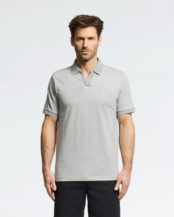 A man in a Psycho Bunny MENS MACKAY JACQUARD JOHNNY COLLAR POLO - B6K408F200, crafted from Pima cotton in light gray with a chest logo, stands against a white background. The polo pairs well with his dark pants. He has short brown hair and a neutral expression.