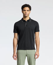 A person is wearing a Psycho Bunny MENS BRYCE PERFORMANCE PIQUE POLO in black, featuring a small chest emblem, paired with light gray pants against a plain white background.