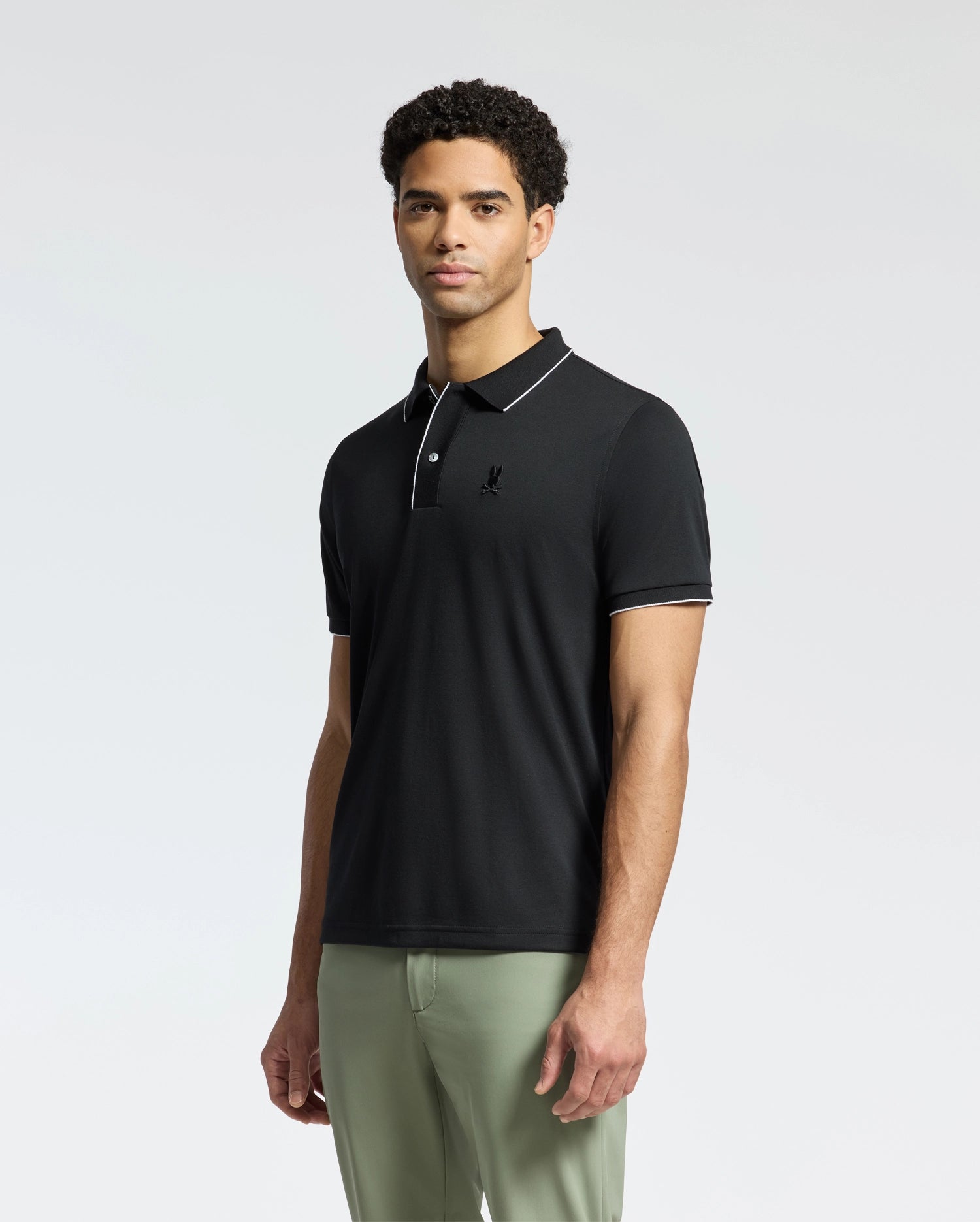 A person with short, curly hair stands against a white background, wearing a Psycho Bunny MENS BRYCE PERFORMANCE PIQUE POLO (B6K486F200) in black with white piping and light green pants. The moisture-wicking polo features a small emblem on the left chest.