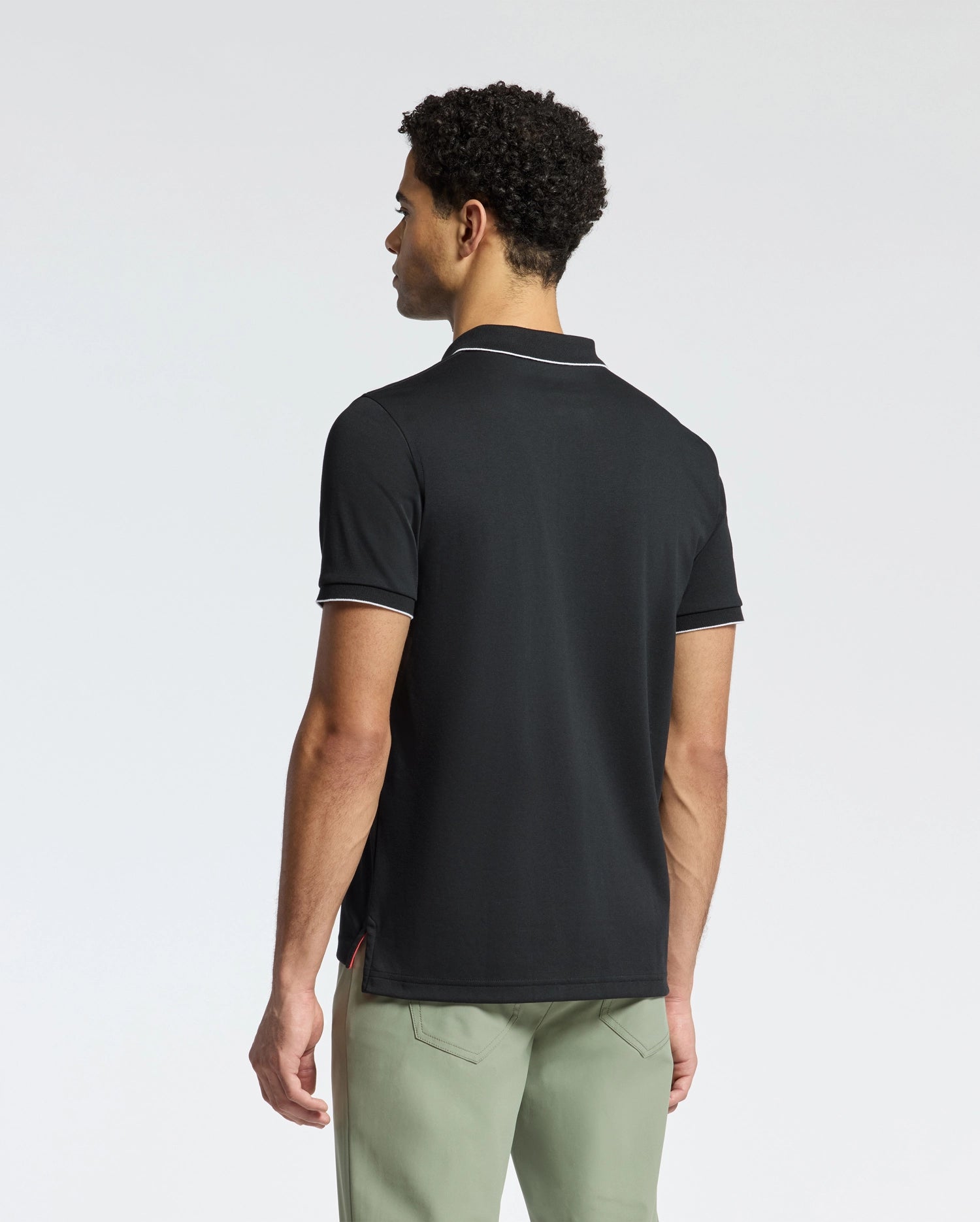 A person with short curly hair, facing away in a plain white background, wears Psycho Bunny's black MENS BRYCE PERFORMANCE PIQUE POLO (B6K486F200) featuring white trim paired with light gray pants.
