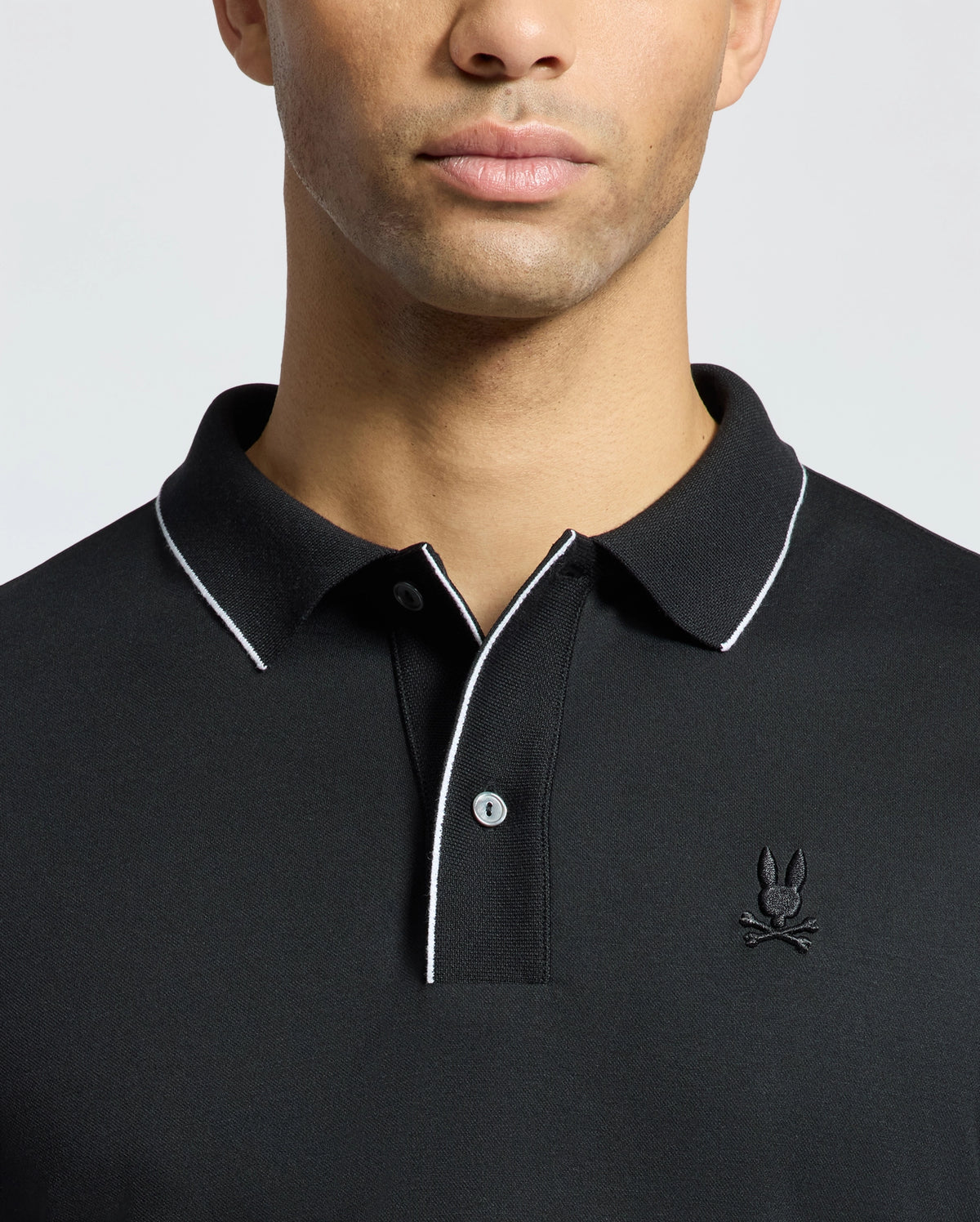 A person wearing the Psycho Bunny MENS BRYCE PERFORMANCE PIQUE POLO, black with white trim on the collar and placket. It features a stylized embroidered logo on the left chest, set against a plain white background.