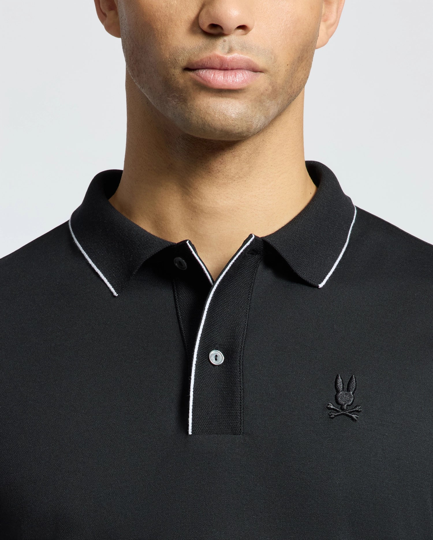 A person wearing the Psycho Bunny MENS BRYCE PERFORMANCE PIQUE POLO, black with white trim on the collar and placket. It features a stylized embroidered logo on the left chest, set against a plain white background.