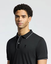 A person in a Psycho Bunny MENS BRYCE PERFORMANCE PIQUE POLO - B6K486F200, black with white trim and logo, stands against a light gray background. The Modal-Pima cotton shirt keeps them cool with moisture-wicking properties as their short, curly hair frames their direct gaze at the camera.