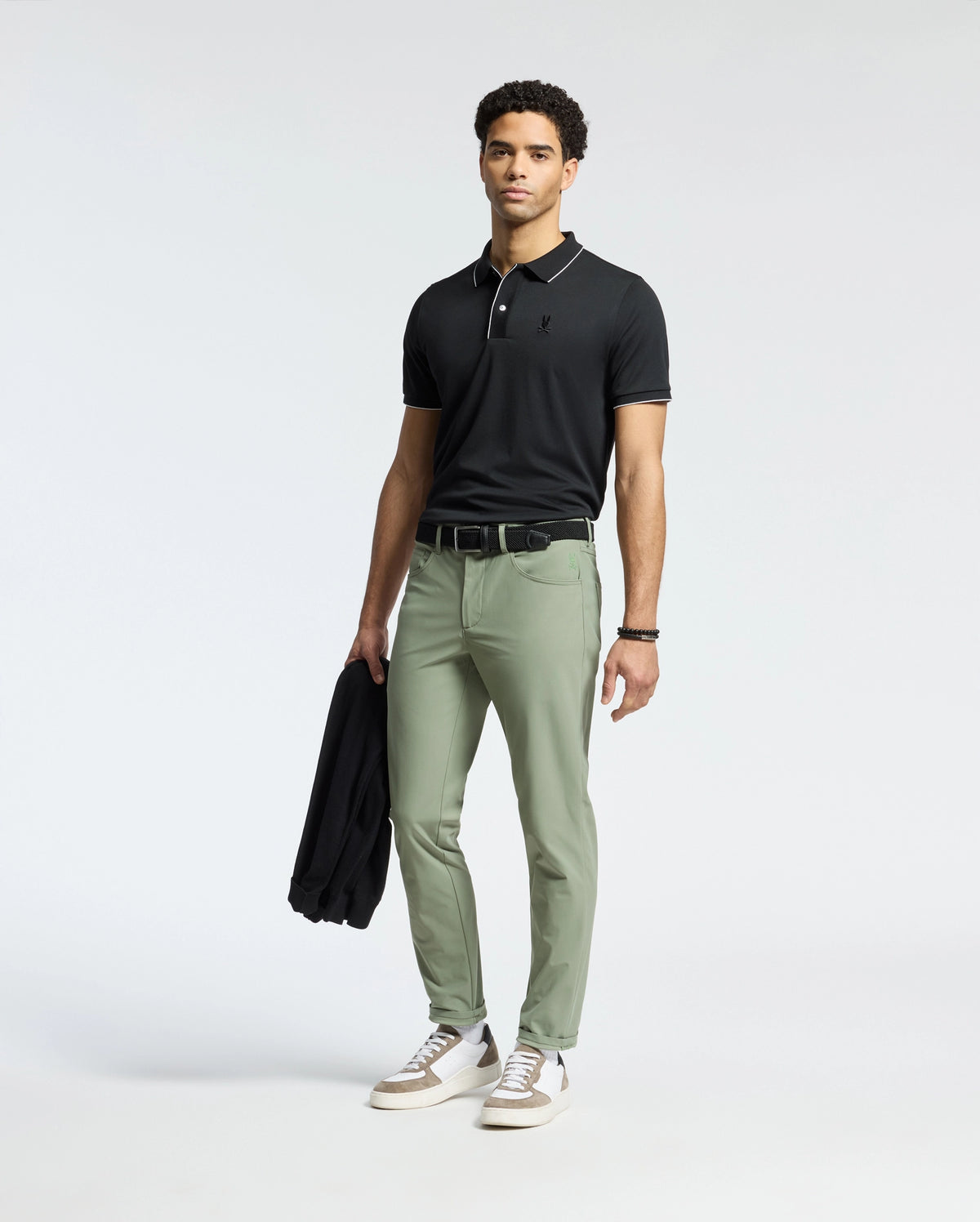 Dressed in a Psycho Bunny MENS BRYCE PERFORMANCE PIQUE POLO, black, with light green pants, the person holds a black jacket. They wear a bracelet on their left wrist and white sneakers with gray and brown accents, standing against a plain white background.