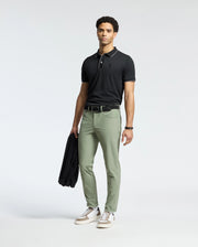 Dressed in a Psycho Bunny MENS BRYCE PERFORMANCE PIQUE POLO, black, with light green pants, the person holds a black jacket. They wear a bracelet on their left wrist and white sneakers with gray and brown accents, standing against a plain white background.