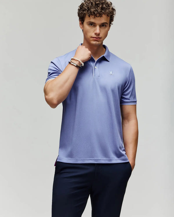 A man with curly hair wears the Psycho Bunny MENS BRYCE PERFORMANCE PIQUE POLO SHIRT (B6K486F200) in light purple and dark pants, standing against a plain light background with one hand in his pocket and the other on his neck.