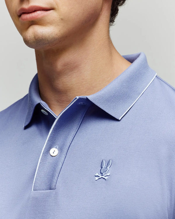 A close-up shows a person in a light blue Psycho Bunny MENS BRYCE PERFORMANCE PIQUE POLO SHIRT, featuring white trim and an embroidered bunny head above crossed bones on the chest. The face is partially visible.