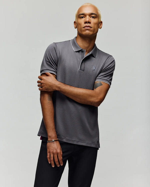 A person with short blond hair models the Psycho Bunny MENS BRYCE PERFORMANCE PIQUE POLO SHIRT - B6K486F200 in gray with white piping, paired with black pants, standing against a plain light background with one arm crossed.