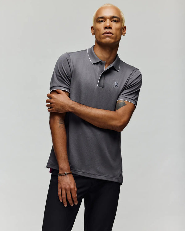 A person with short blond hair models the Psycho Bunny MENS BRYCE PERFORMANCE PIQUE POLO SHIRT - B6K486F200 in gray with white piping, paired with black pants, standing against a plain light background with one arm crossed.