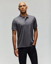 A man with short blond hair wears the Psycho Bunny MENS BRYCE PERFORMANCE PIQUE POLO SHIRT (B6K486F200) in gray, featuring a small embroidered logo, paired with black pants. He stands against a plain light background, one hand in his pocket.
