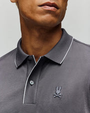 A man is seen from the lower face to upper torso wearing the Psycho Bunny MENS BRYCE PERFORMANCE PIQUE POLO SHIRT (B6K486F200), a dark gray polo with white collar trim and an embroidered bunny and crossbones logo on the chest.