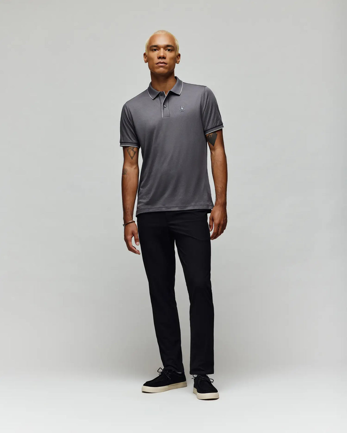 A person with short blond hair stands against a plain light gray background, wearing the Psycho Bunny MENS BRYCE PERFORMANCE PIQUE POLO SHIRT (B6K486F200), black pants, and black shoes with white soles.