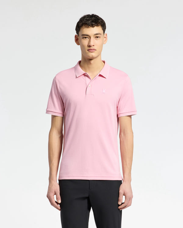 A person stands against a white backdrop wearing Psycho Bunny's Men's Bryce Performance Pique Polo in light pink, made from performance pique fabric with a small chest logo. They have short dark hair and wear black quick-dry, moisture-wicking pants.