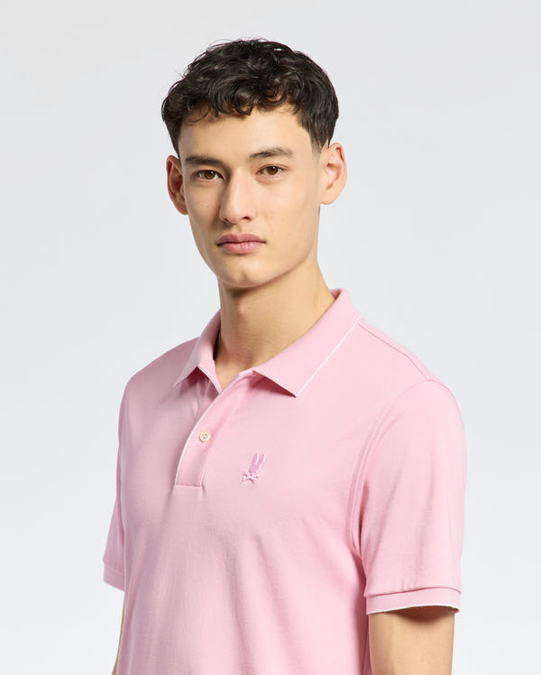 A person wearing the Psycho Bunny MENS BRYCE PERFORMANCE PIQUE POLO in light pink, featuring a small logo on the chest, stands against a plain background. They have short, dark hair and are looking directly at the camera.