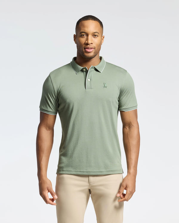 A man stands against a plain background wearing the Psycho Bunny MENS BRYCE PERFORMANCE PIQUE POLO (B6K486F200) in light green, made from breathable moisture-wicking fabric with a small embroidered logo on the chest, paired with beige pants.