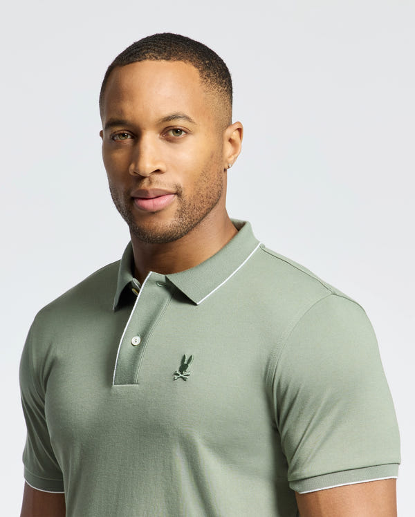 A person wears the Psycho Bunny Men's Bryce Performance Pique Polo (B6K486F200) in light green, made from breathable, moisture-wicking Modal-Pima cotton. A small dark green logo adorns the chest as they stand facing forward against a plain, softly lit backdrop.