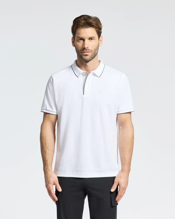 A man stands against a plain background wearing the Psycho Bunny MENS BRYCE PERFORMANCE PIQUE POLO (B6K486F200) in white with black trim and black pants, designed for ultimate comfort and moisture-wicking.