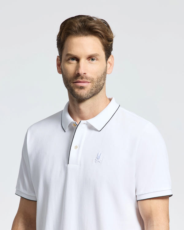 A man with short brown hair and a beard is wearing the Psycho Bunny MENS BRYCE PERFORMANCE PIQUE POLO - B6K486F200 in white, designed for moisture-wicking. He stands against a plain light gray background, looking directly at the camera with a neutral expression.