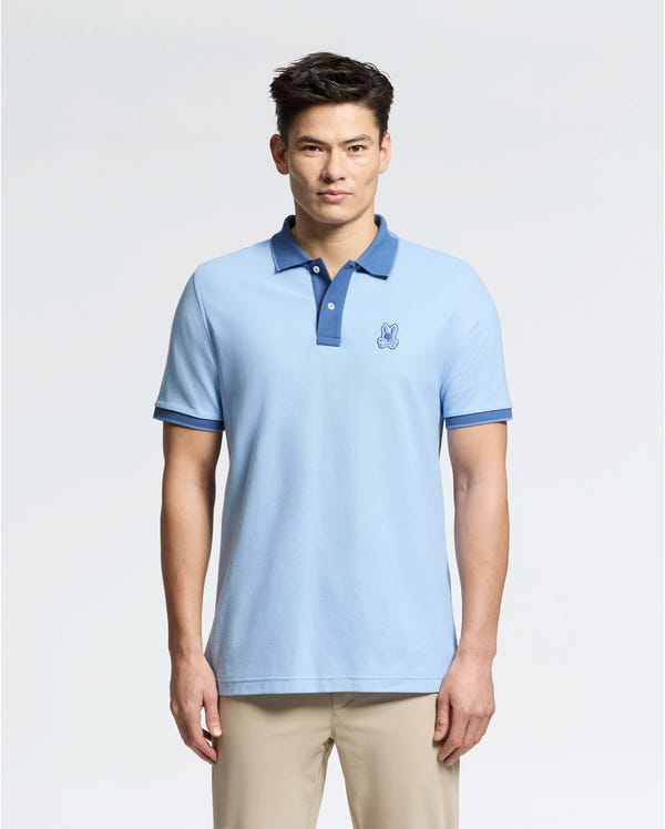 A man wears a Psycho Bunny slim-fit light blue "MENS NEW CASTLE PIQUE POLO - B6K487F200" with an embroidered bunny patch, dark blue collar, and sleeve accents, against a plain background. It's stylishly paired with beige pants.