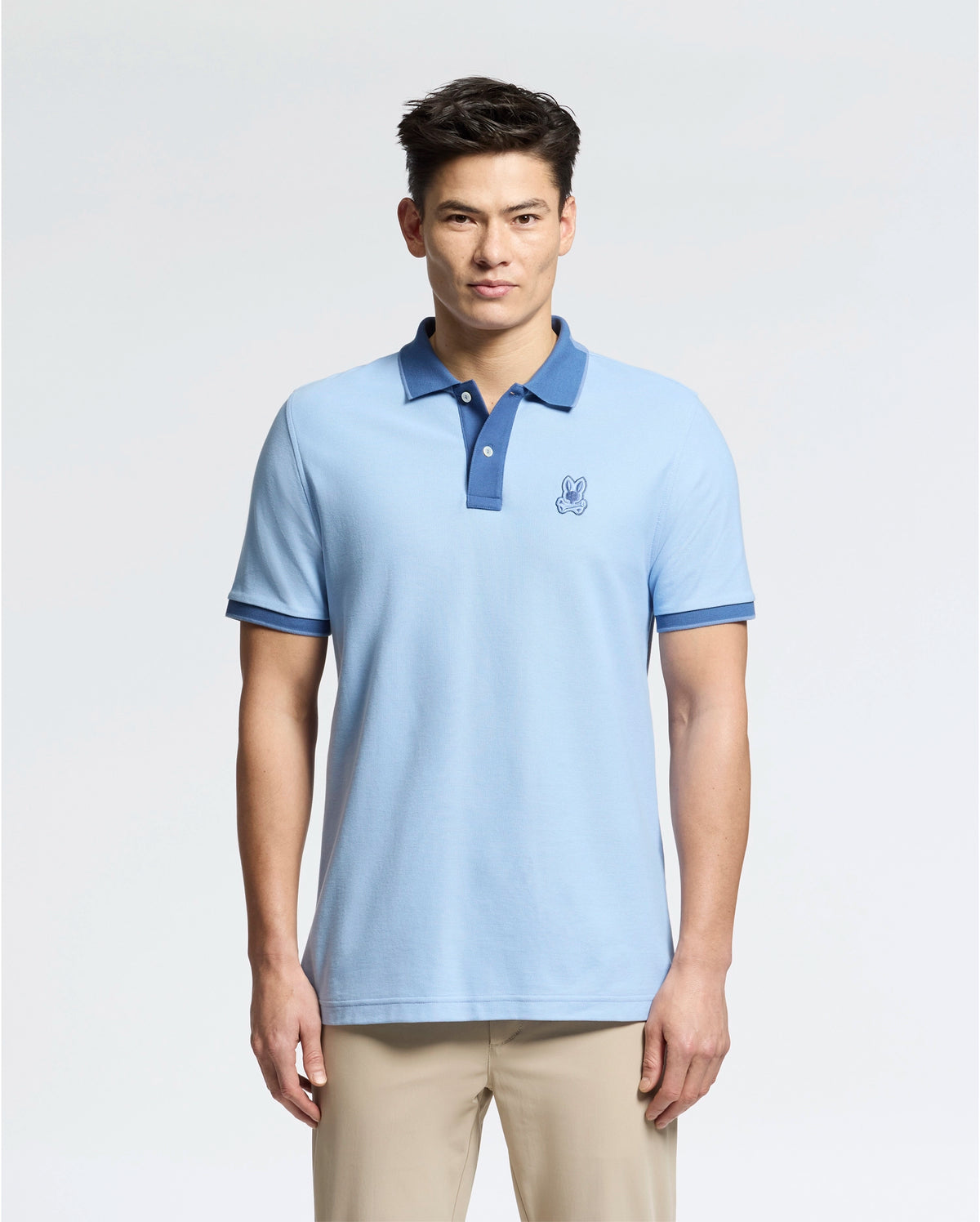A man wears a Psycho Bunny slim-fit light blue "MENS NEW CASTLE PIQUE POLO - B6K487F200" with an embroidered bunny patch, dark blue collar, and sleeve accents, against a plain background. It's stylishly paired with beige pants.